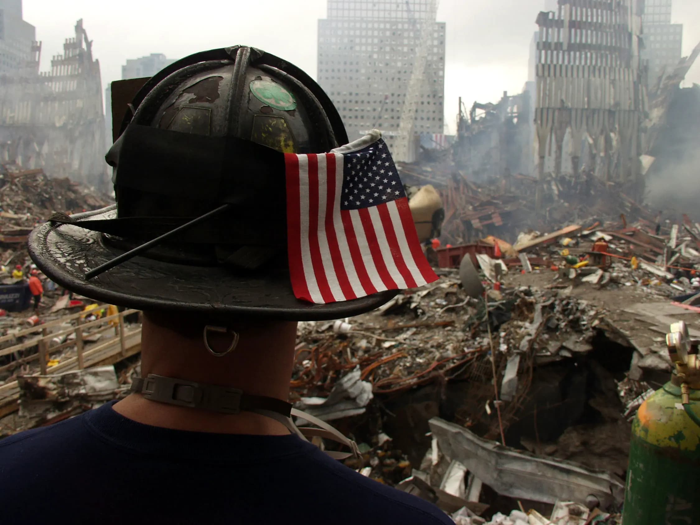 A firefighter wearing a black helmet with a small American flag tucked into it stands with their back to the camera, surveying a vast field of rubble and debris